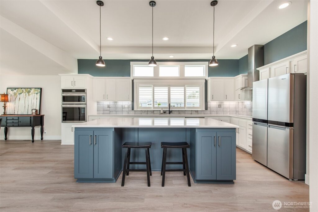 A modern, open-concept kitchen featuring a large central island with a white countertop and a dusty blue base, flanked by two black wooden barstools