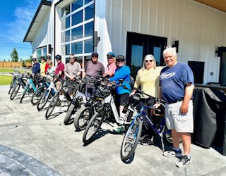 A group of about ten senior cyclists standing outside a modern white building with their bicycles on a sunny day