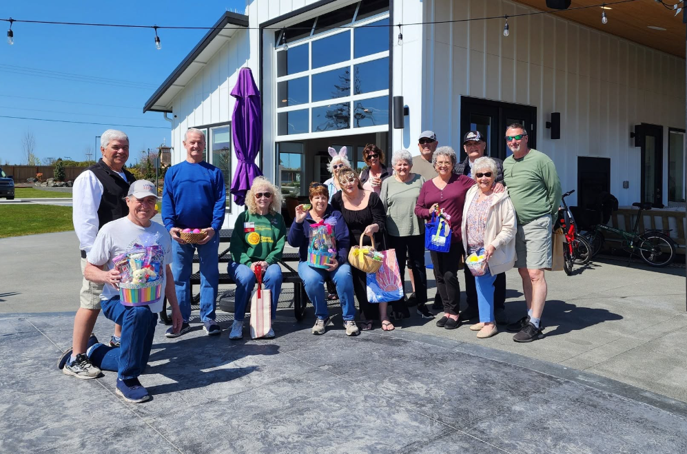 A group of about fifteen smiling adults posing together outside a modern clubhouse on a clear, sunny day